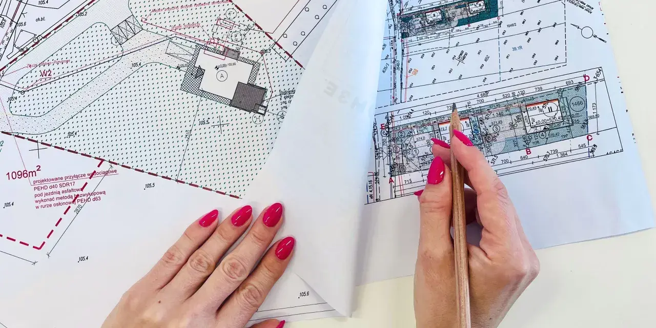Dłoń z różowym lakierem na paznokciach trzymająca ołówek nad mapą projektową.