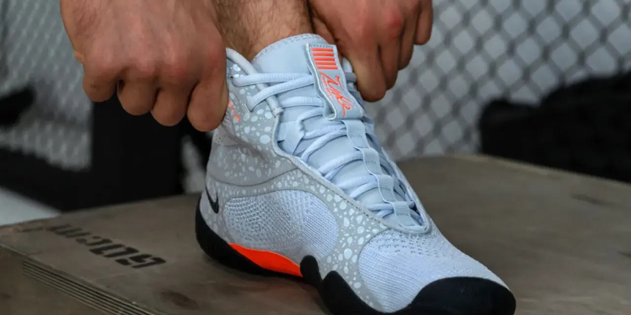 Ręce zapinają jasnoniebieskie buty sportowe z pomarańczowymi akcentami, przypominające styl Batistini.