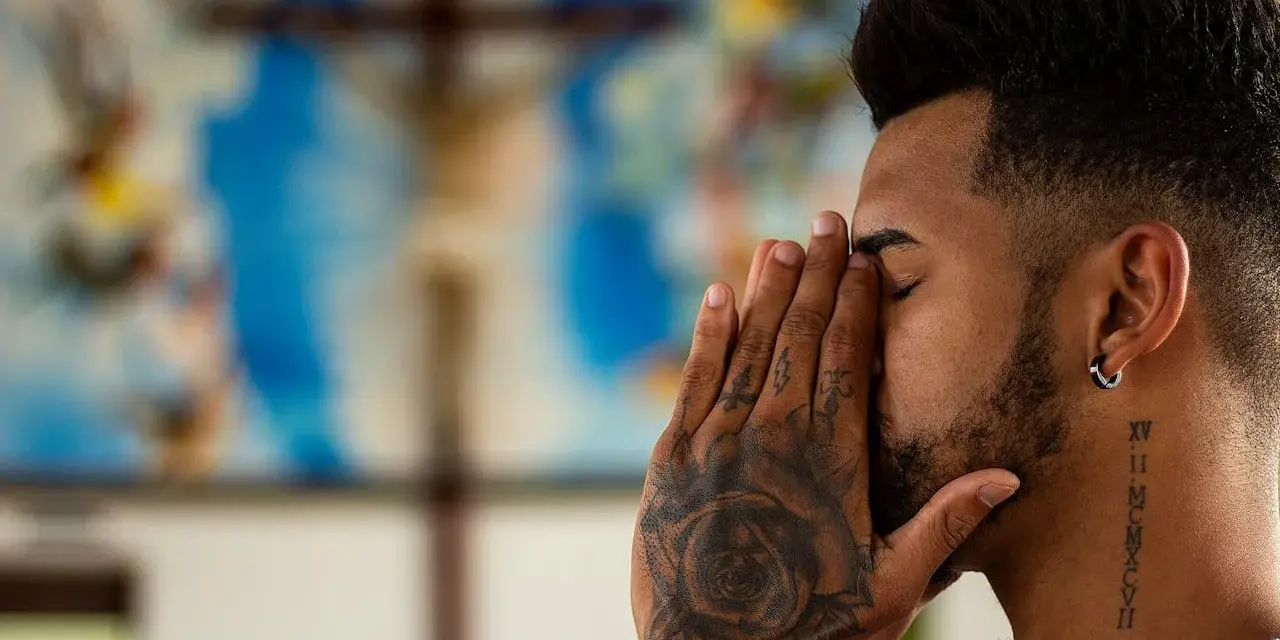 A man with tattoos prays before a crucifix, contemplating if is vaping a sin.