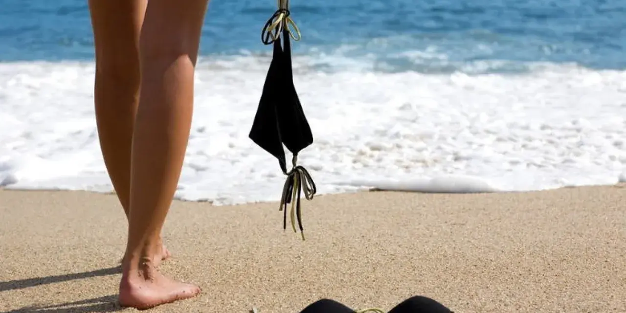 Nogi na piaszczystej plaży, obok czarny kostium kąpielowy. Fale obmywają brzeg. Idealne miejsce na naturystyczną plażę.