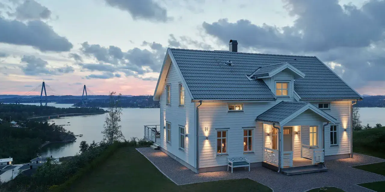 Vackert vitt hus med upplysta fönster och entré, perfekt hus entre inspiration. Vacker vy över vatten och bro i skymningen.