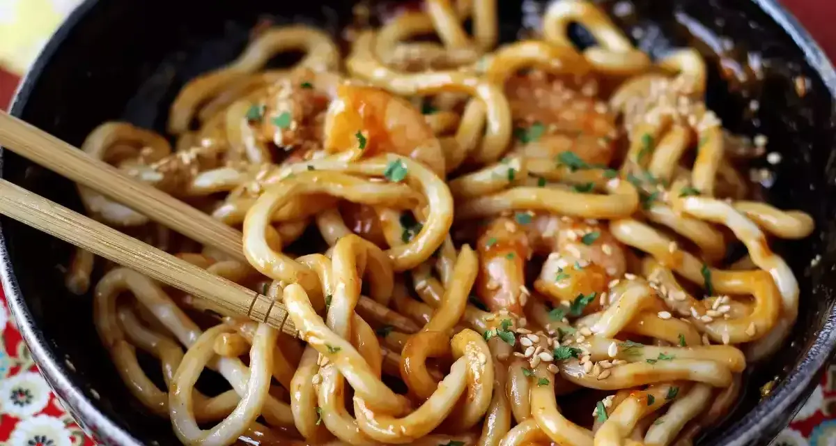 Miseczka z makaronem udon z krewetkami, posypana sezamem i pietruszką. Pałeczki zanurzone w daniu. Prosty przepis na to, jak gotować makaron udon.