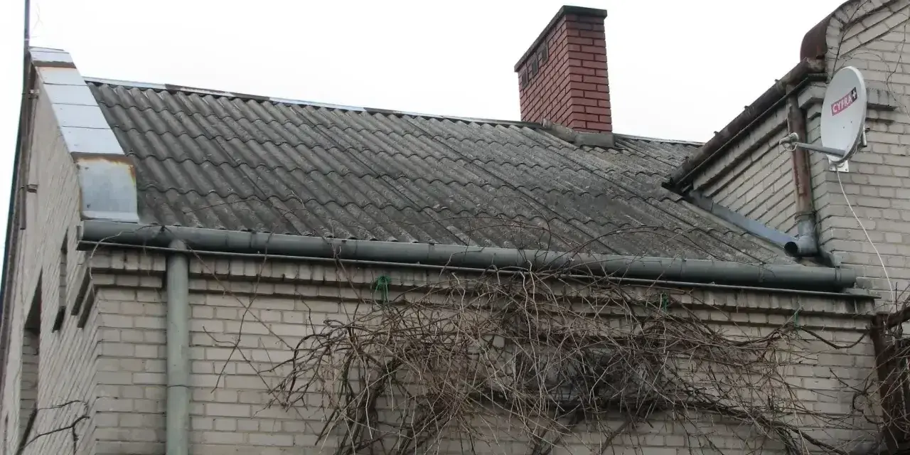 Dach pokryty eternitem, który zawiera azbest. Co to za materiał? Stary, szary eternit na dachu domu, z kominem i anteną satelitarną.