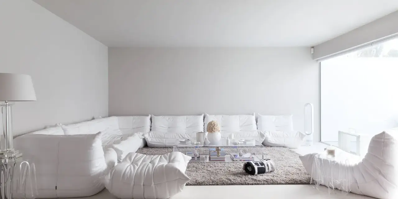 Soggiorno minimalista con divano componibile bianco, tappeto grigio e tavolino trasparente. Le tonalità di bianco per pareti creano un'atmosfera eterea.