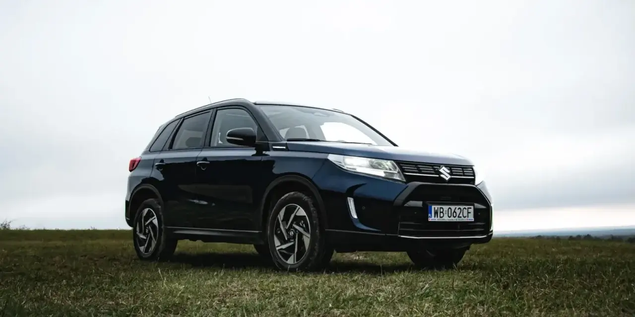 Ciemnoniebieski Suzuki Vitara Hybrid na tle zachmurzonego nieba, zaparkowany na trawiastym wzgórzu.