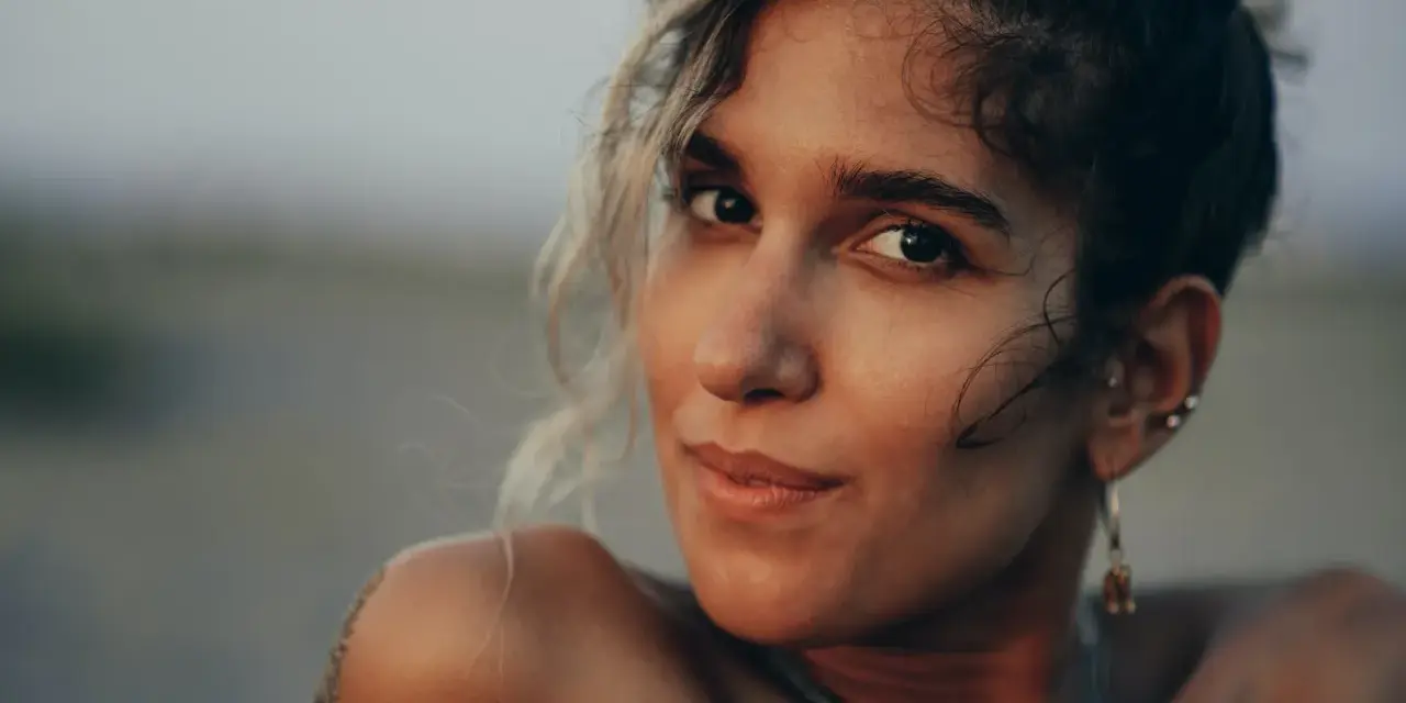 Photos de portrait d'une jeune femme aux cheveux bouclés, le sable sur son épaule, regardant la caméra avec un léger sourire.