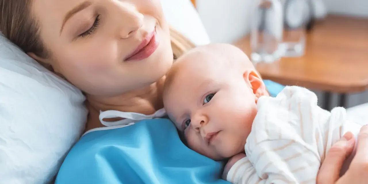 Uśmiechnięta mama tuli noworodka. To chwila spokoju po szczepieniach noworodka, budująca więź i zapewniająca zdrowie maluszka.