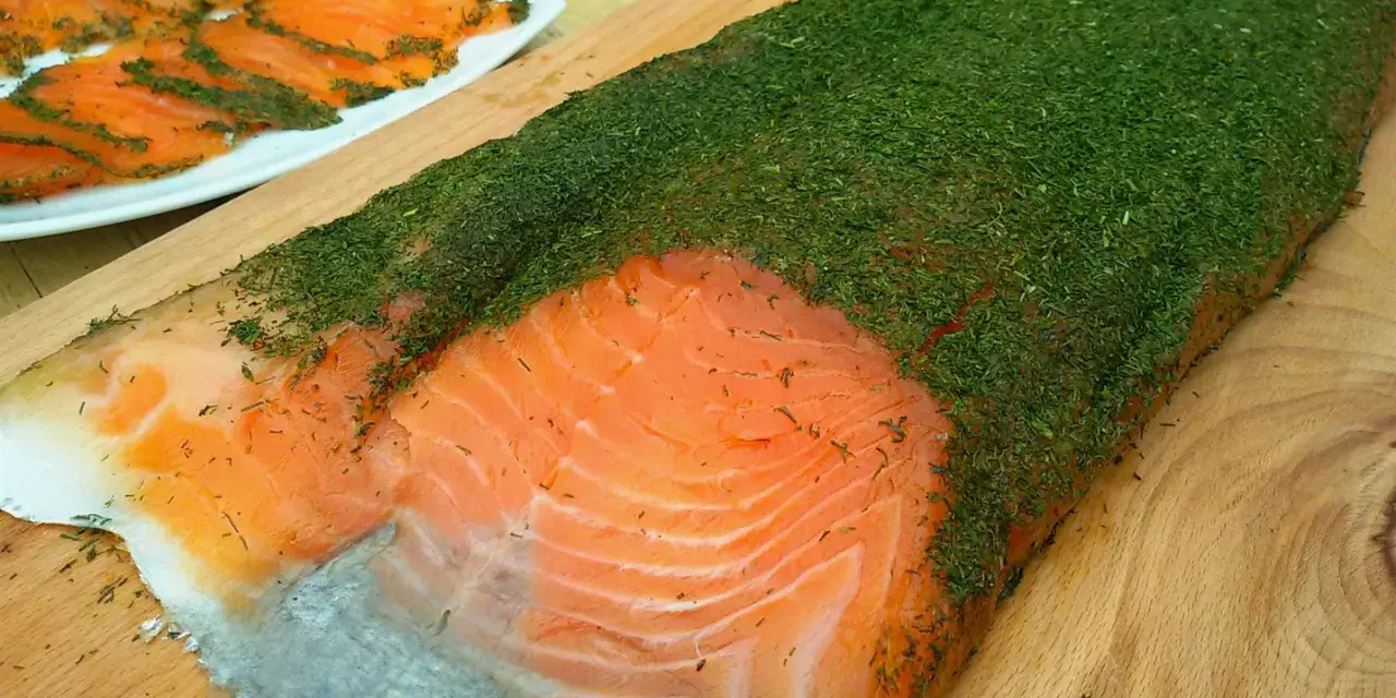 Un gran filete de salmón marinado casero cubierto de eneldo fresco. En el fondo, finas lonchas de salmón ya preparadas.
