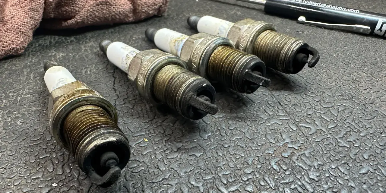 Four used spark plugs lie on a textured surface next to a rag and a screwdriver. This image might make you wonder how many spark plugs a car has.
