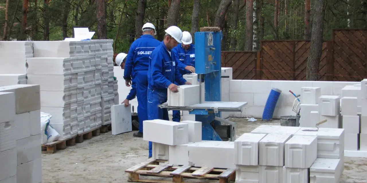 Budowa trwa! Pracownicy w niebieskich kombinezonach i białych kaskach układają bloczki ytong. Na paletach widać, że ytong 12 to spora ilość materiału budowlanego.