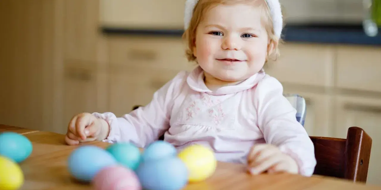 Kleines Mädchen mit Hasenohren und bunten Ostereiern. Sie sitzt am Tisch und schaut neugierig auf das **Baby Ei**.