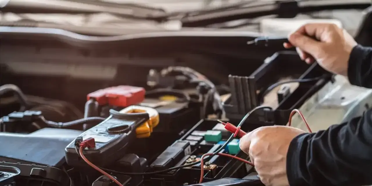 Mechanik sprawdza instalację elektryczną samochodu. Na zdjęciu widać multimetr i ręce trzymające sondy. Stojan z bezpiecznikami jest widoczny w tle.
