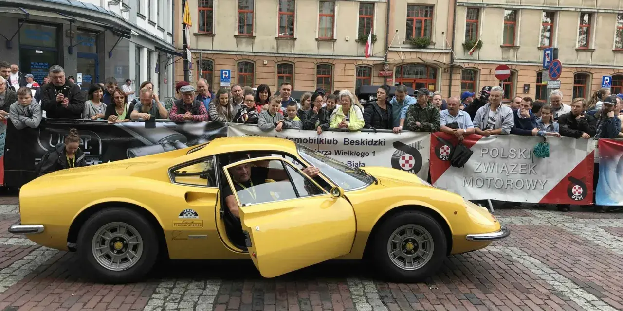 Żółte Ferrari Dino z otwartymi drzwiami, kierowca w środku, tłum widzów za barierką.