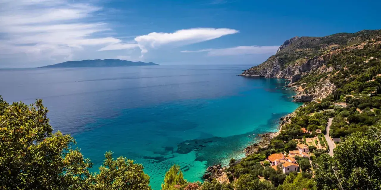 Turquoise water laps a rocky Tuscan coast with villas nestled among trees, under a blue sky with clouds.
