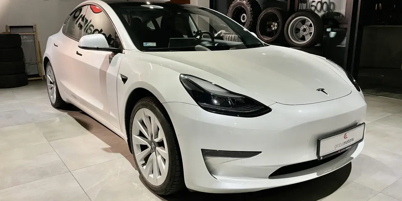 Biała Tesla Model 3 z napędem na cztery koła stoi w salonie samochodowym.