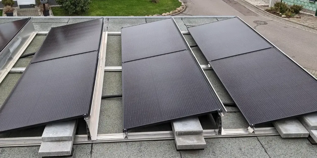 Solarmodule auf einem Bitumen-Flachdach, befestigt mit einer Aufständerung. Ein kleiner schwarzer Kater beobachtet die Installation.