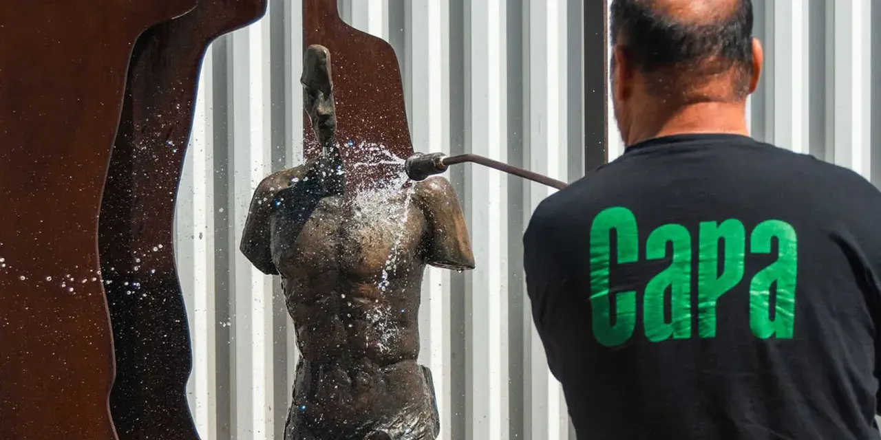 Hombre con camiseta negra "Capa" limpia una escultura de bronce con agua a presión.