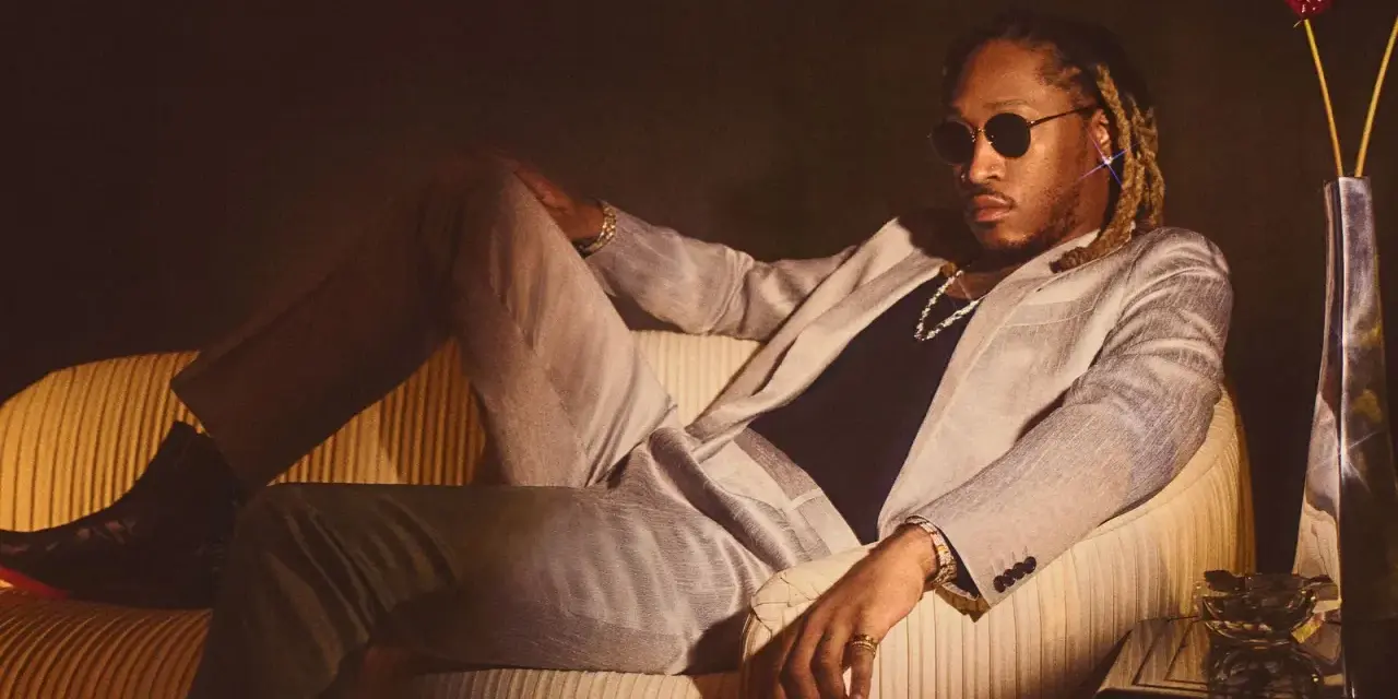 Ein Hip-Hop-Künstler in einem schicken Anzug entspannt sich auf einem Sofa, trägt Sonnenbrille und Schmuck.