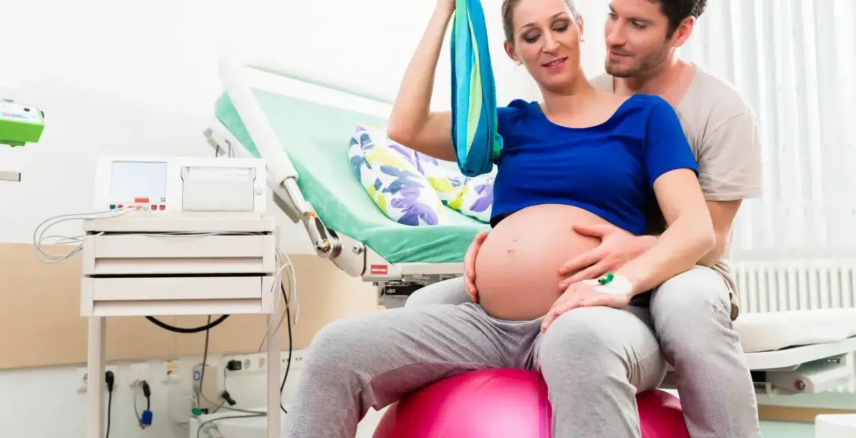 Para z niecierpliwością oczekuje porodu. Kobieta siedzi na piłce porodowej, wspierając się na chuście. Mężczyzna obejmuje ją czule. To może być ich pierwszy poród, ale przygotowują się na VBAC.