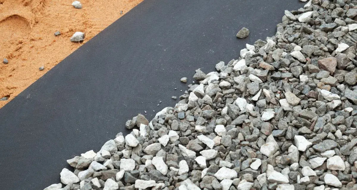 Gros plan sur un géotextile noir recouvert de gravier gris et blanc, à côté de sable. Idéal pour estimer le prix pose géotextile et gravier.