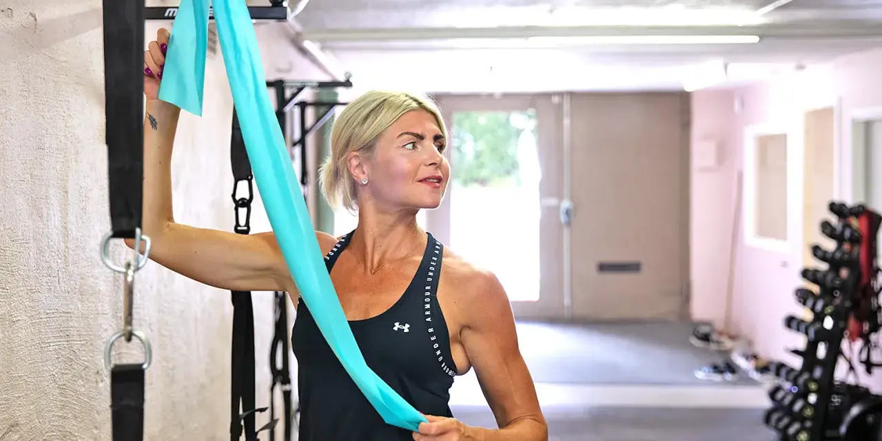 Kvinna med blont hår stretchar axlar med ett turkost träningsband i ett gym.