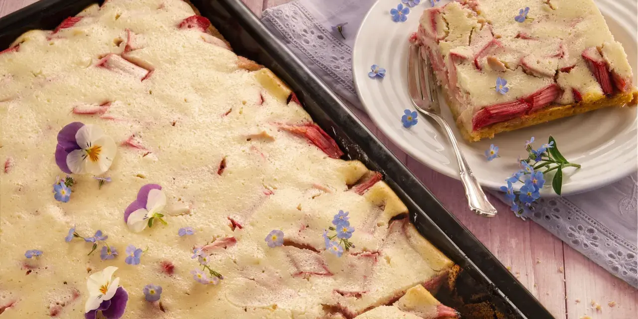 Saftiger Rhabarberkuchen mit Quark, dekoriert mit Veilchen und Vergissmeinnicht. Ein Stück ist auf einem Teller angerichtet.
