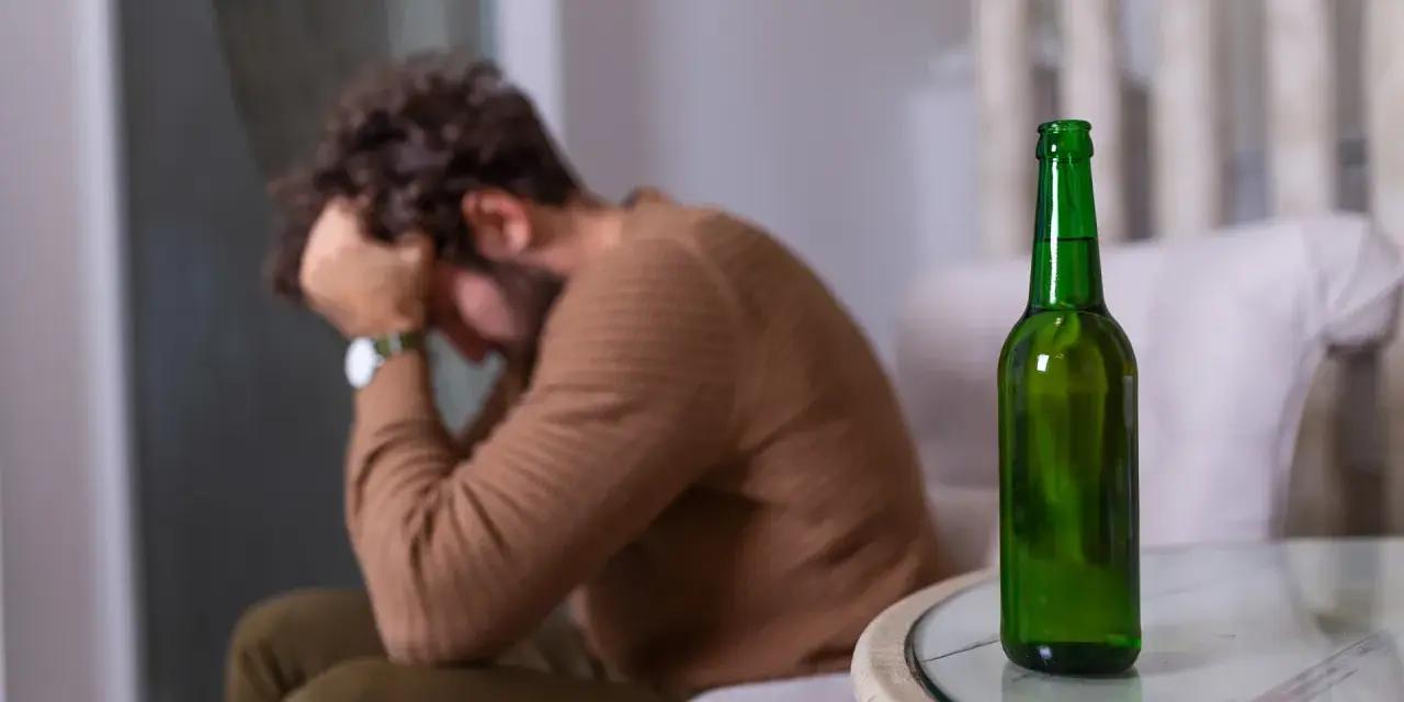 Mężczyzna siedzi z głową w dłoniach, obok stoi zielona butelka. Czuje duszności po alkoholu.