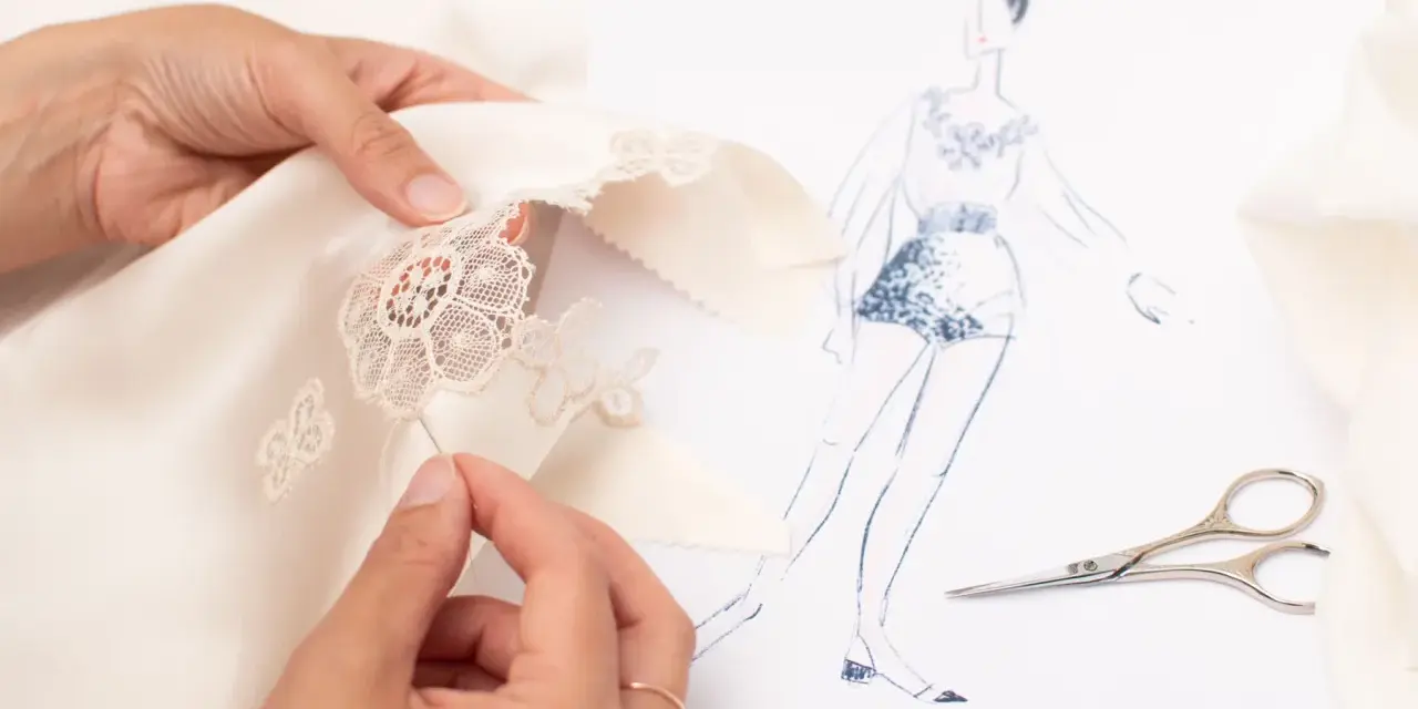 Main d'une couturière réalisant un assemblage couture délicat sur un tissu de soie, avec un croquis de mode et des ciseaux à proximité.