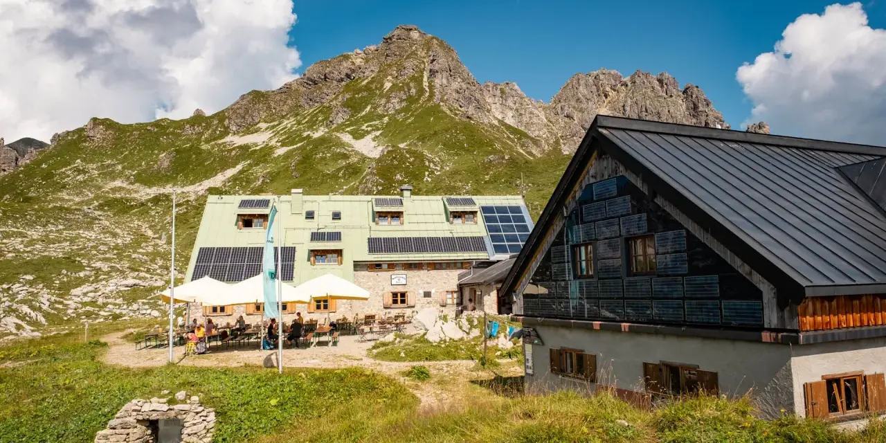 Die Buchel Alpe, eine Berghütte mit Solaranlagen, liegt vor einer beeindruckenden Bergkulisse. Gäste sitzen draußen unter Sonnenschirmen.
