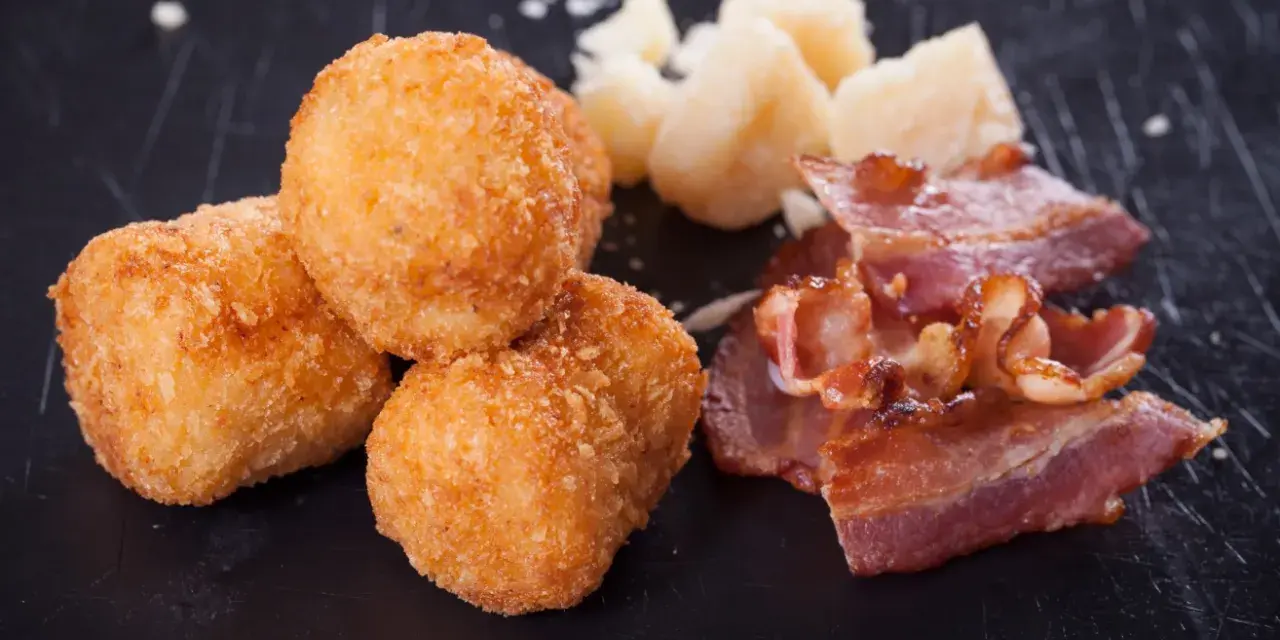 Croquetas doradas y crujientes con trozos de bacon y queso sobre fondo oscuro.
