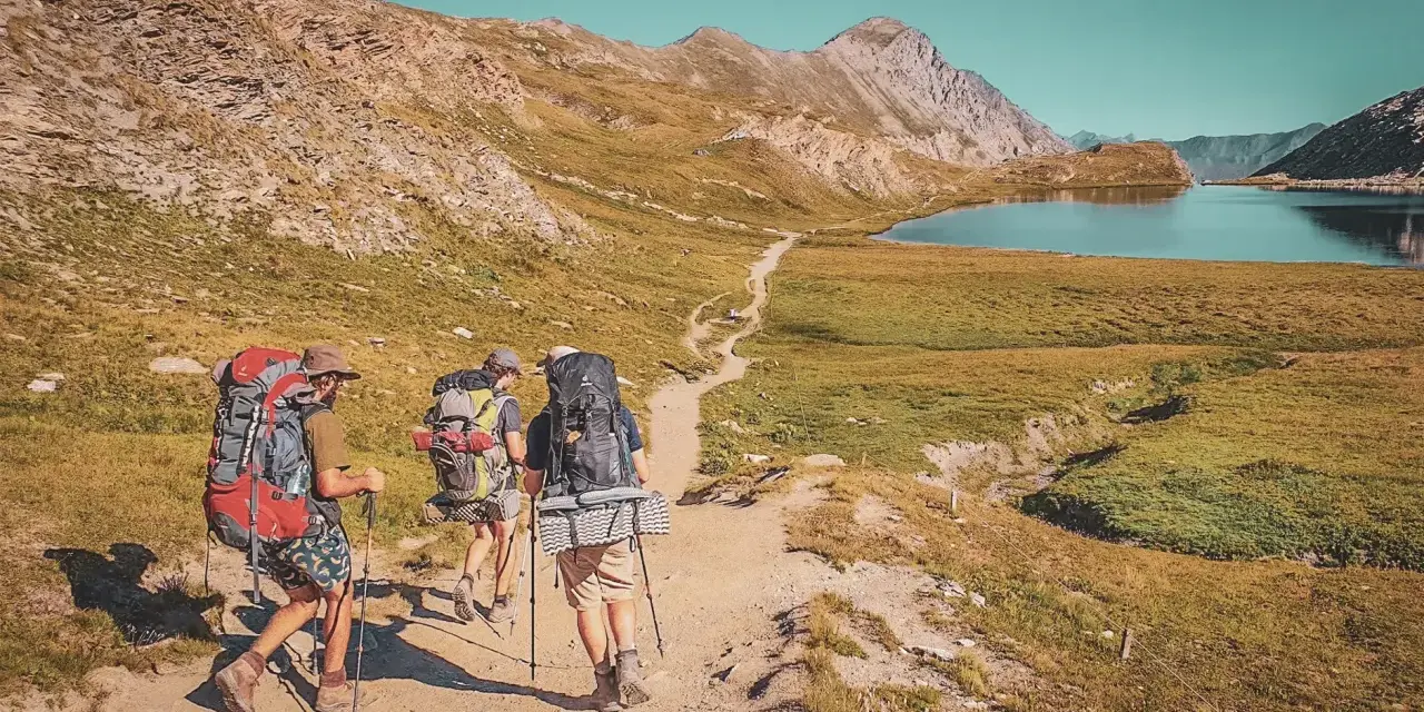 Trois randonneurs équipés pour le tour du Queyras traversent un sentier de montagne, un lac bleu et des sommets rocheux en toile de fond.