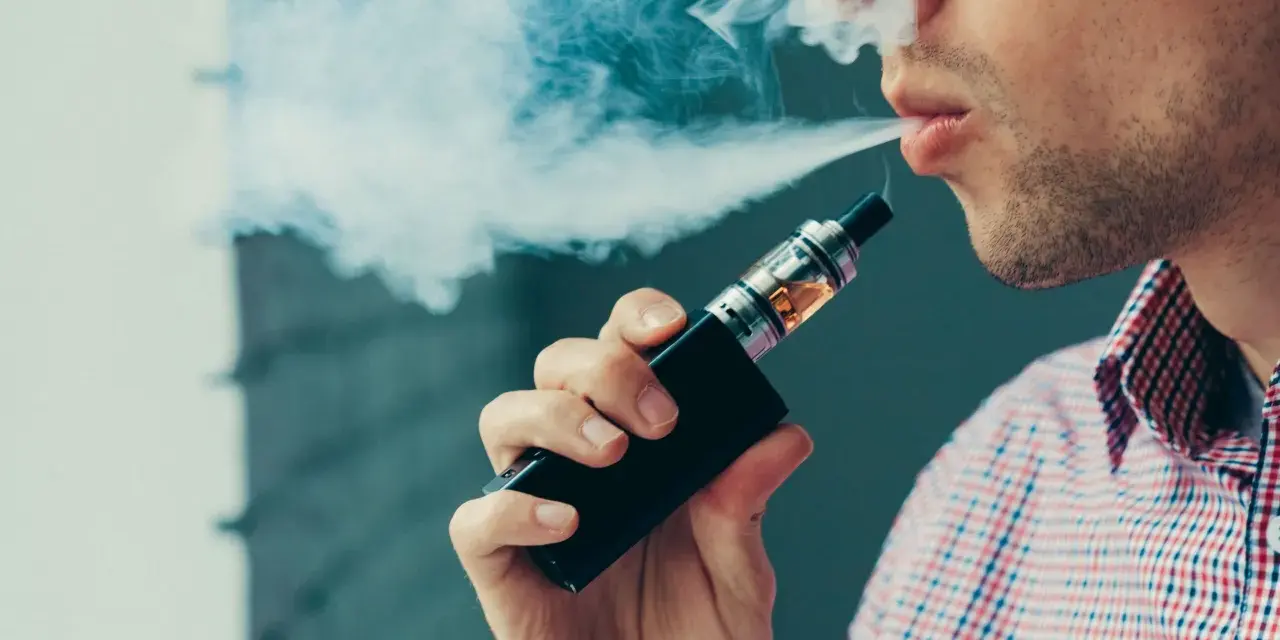 Hombre vapeando, exhala humo blanco. El vapear sube la tensión, cuidado con tu salud.