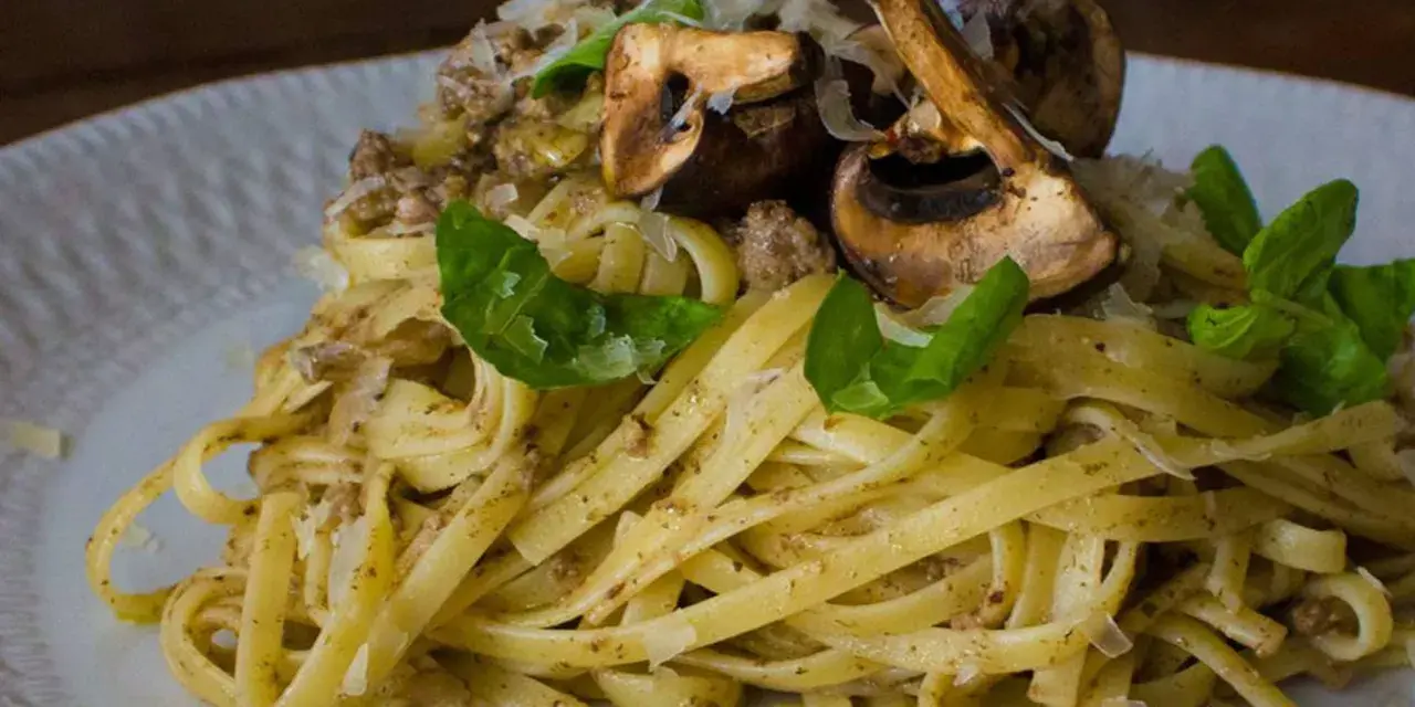 Pasta con champiñones, carne picada y queso rallado, decorada con hojas verdes.