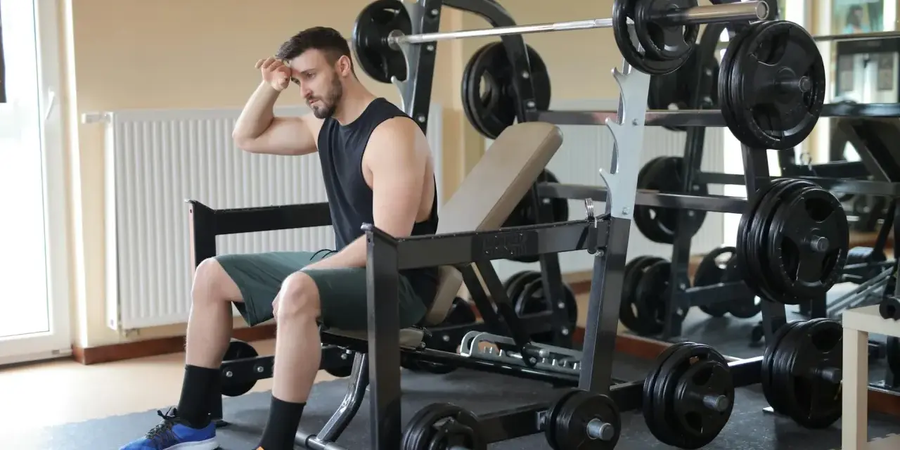 Młody mężczyzna, pierwszy raz na siłowni, siedzi zmęczony na ławce, wycierając pot z czoła. Wokół niego sprzęt do ćwiczeń.