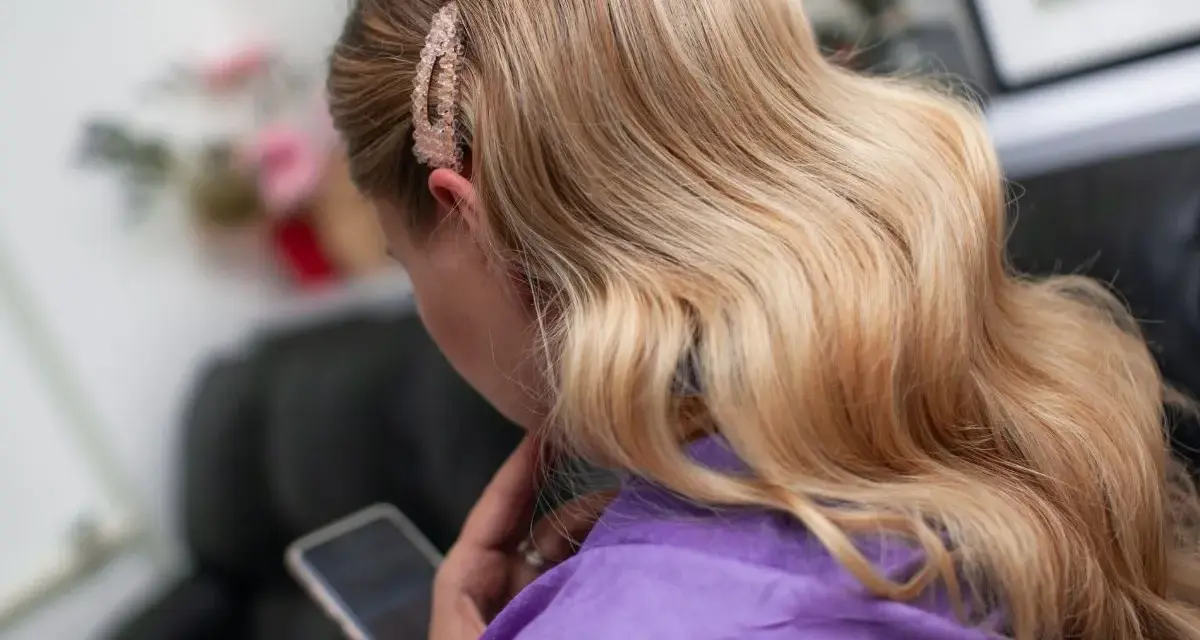 Blond włosy ułożone w hollywoodzkie fale na bok, ozdobione błyszczącą spinką. Kobieta w fioletowym szlafroku przegląda telefon.