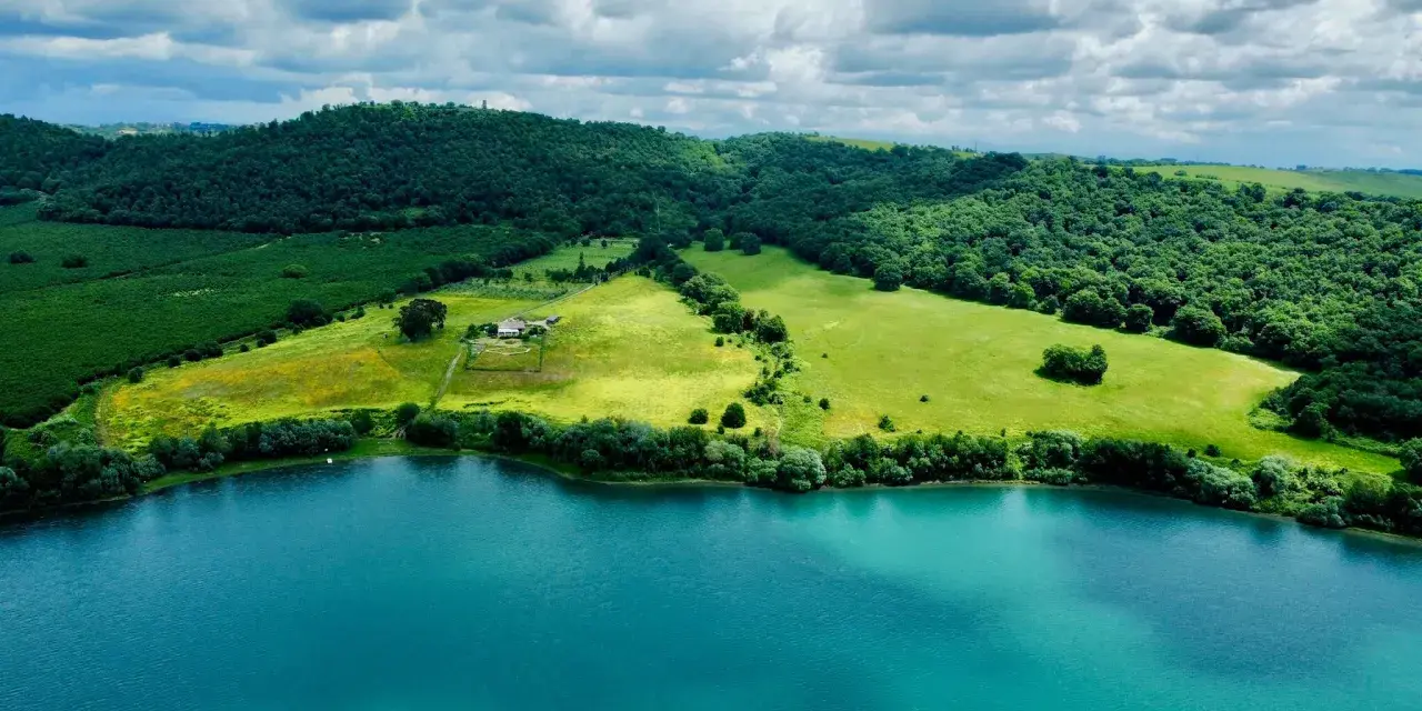 Acque turchesi del lago di Martignano lambiscono dolci colline verdi, con un casale isolato tra i campi.