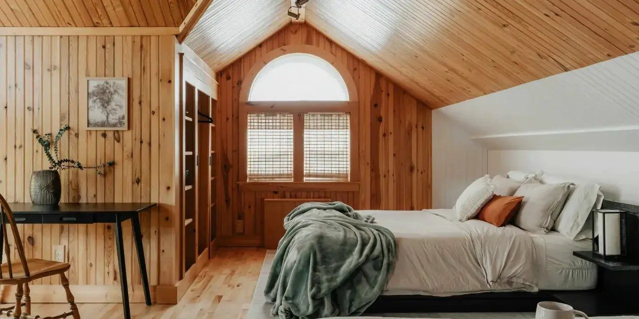 Dormitorio con paredes y techo de madera, cama con mantas y almohadas, ventana arqueada y escritorio.