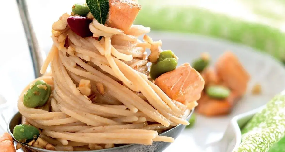 Spaghetti di riso con salmone, fave e noci, guarniti con una foglia verde. Un piatto invitante e colorato.