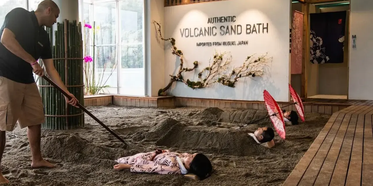A man rakes sand for a volcanic sand bath. Two people lie buried in the sand, with red umbrellas over their heads.