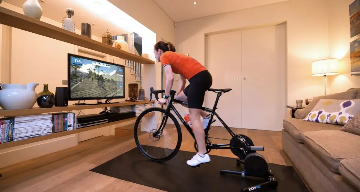 Woman in her home cycling pain cave, training indoors on a bike trainer while watching a virtual race on a large screen.