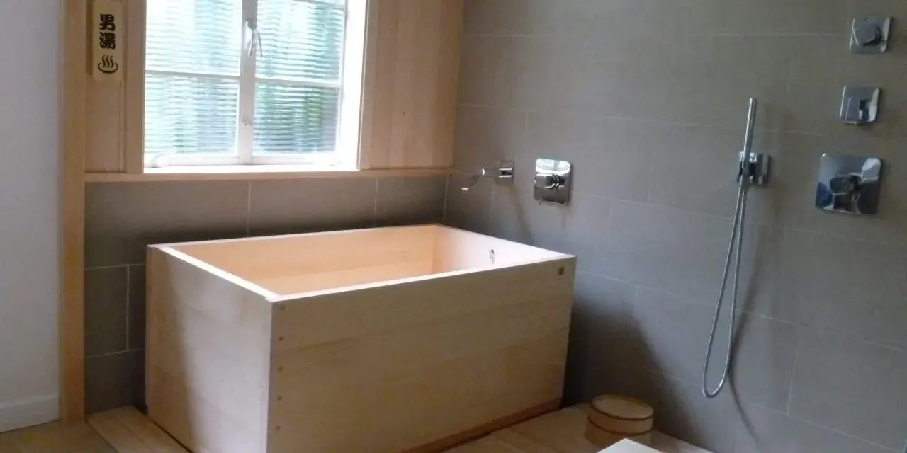 Un baño japonés con tina de madera, ducha y un pequeño taburete. El letrero de madera indica "男湯" (baño de hombres).