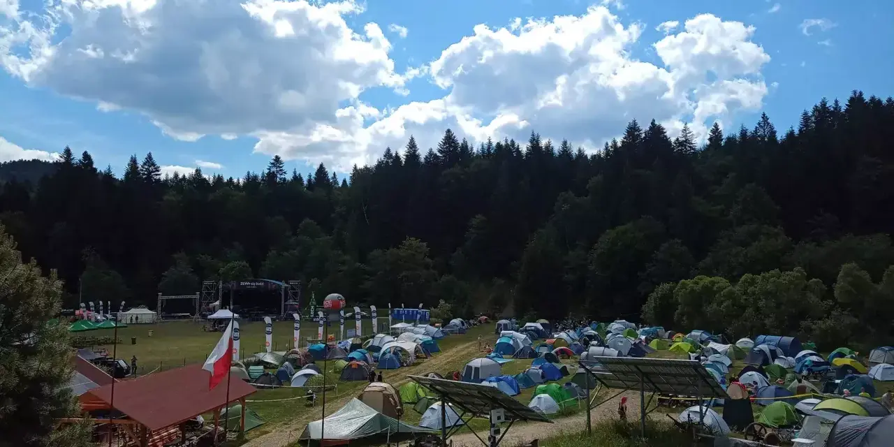 Pole namiotowe na festiwalu w Solinie. Tysiące namiotów pod błękitnym niebem z chmurami, w tle las i scena festiwalowa.