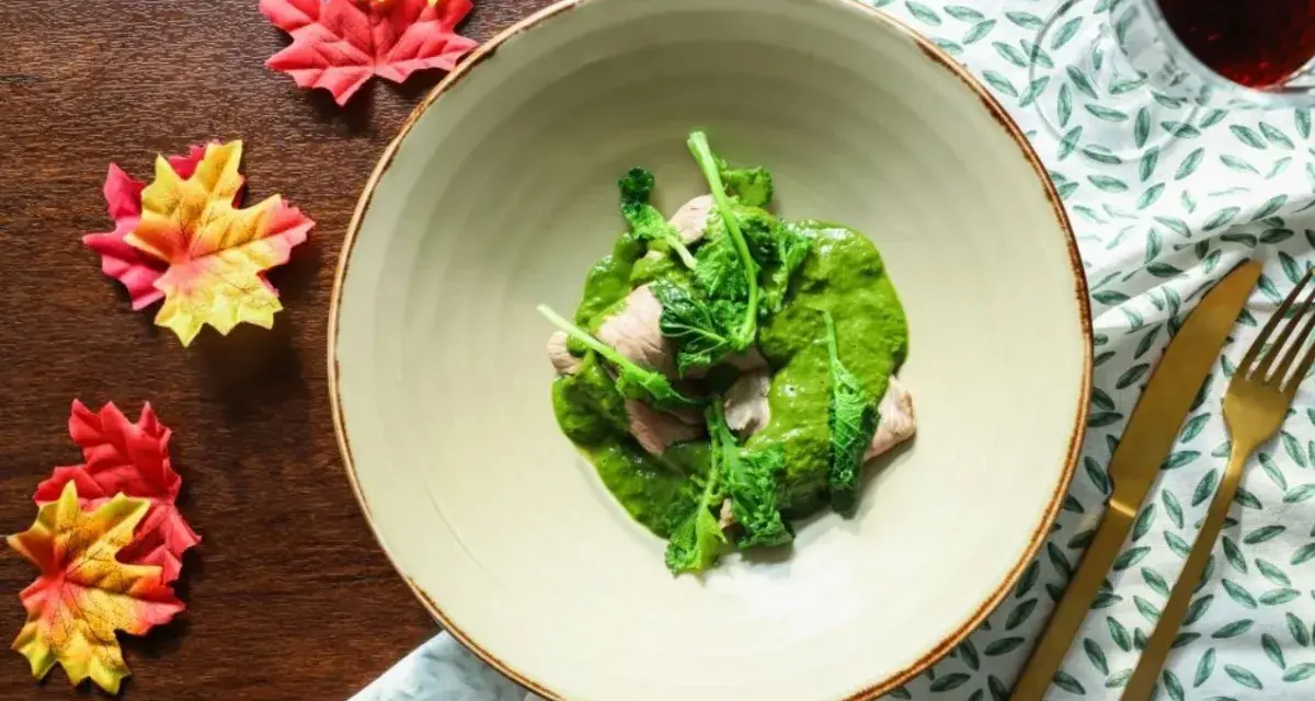 Piatto di carne con salsa verde cremosa, guarnito con cime di rapa fresche. Accanto, foglie autunnali e posate dorate.