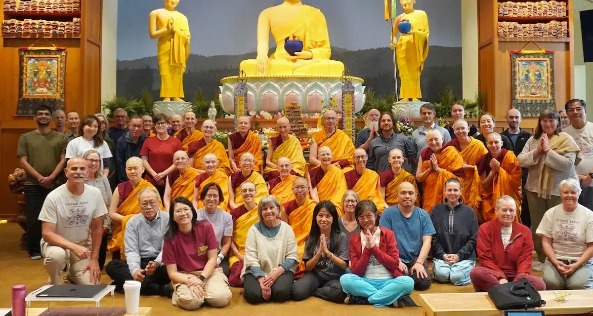 A large group of monks and laypeople gather for a photo in front of a golden Buddha statue, celebrating their shared religion.