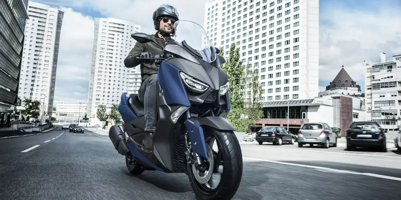 Mężczyzna w kasku i okularach przeciwsłonecznych jedzie niebieskim skuterem Yamaha X-MAX 400 po miejskiej drodze. W tle wysokie budynki i samochody.