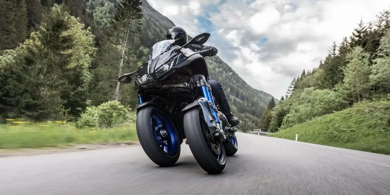 Motocyklista na Yamaha Niken pokonuje zakręt na górskiej drodze, otoczony zielenią i chmurami.