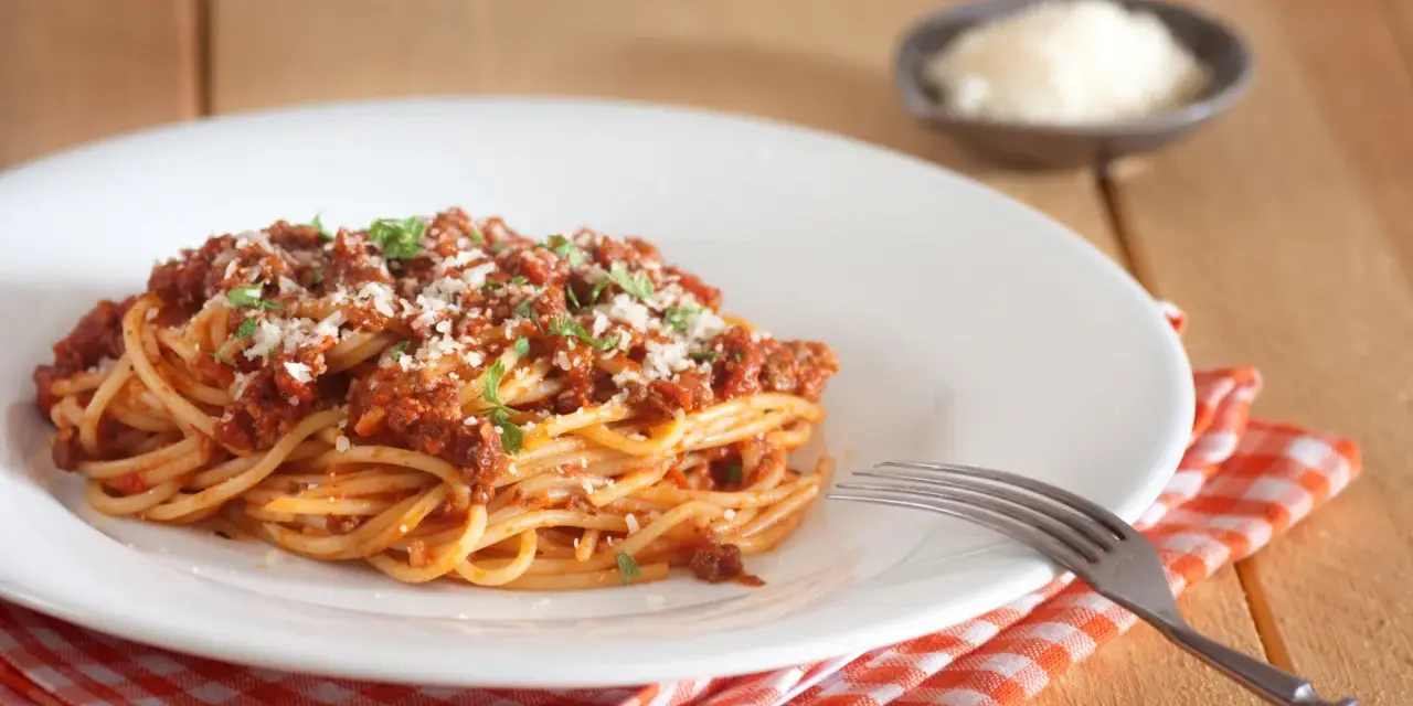 Talerz spaghetti bolognese z parmezanem i natką pietruszki, obok widelec i ser w miseczce.