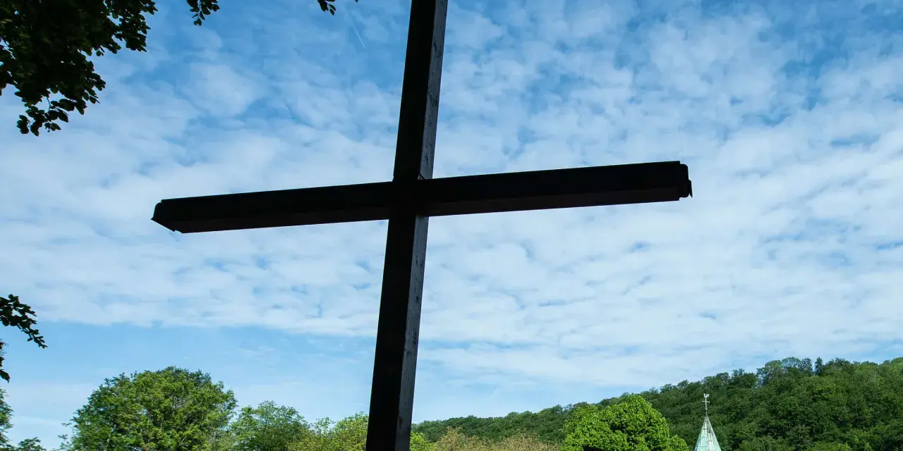 Ein großes Holzkreuz ragt vor blauem Himmel mit Wolken auf. Im Hintergrund eine Kirche und grüne Bäume. Wann ist Christi Himmelfahrt?