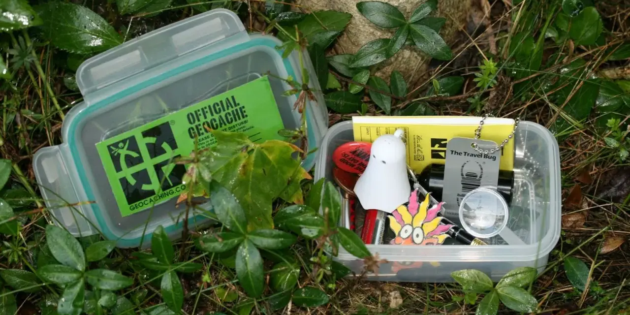 Dwa pojemniki z zawartością do geocaching co to. W jednym zielona naklejka "Official Geocache", w drugim drobne przedmioty jak duch, lupa i "Travel Bug".