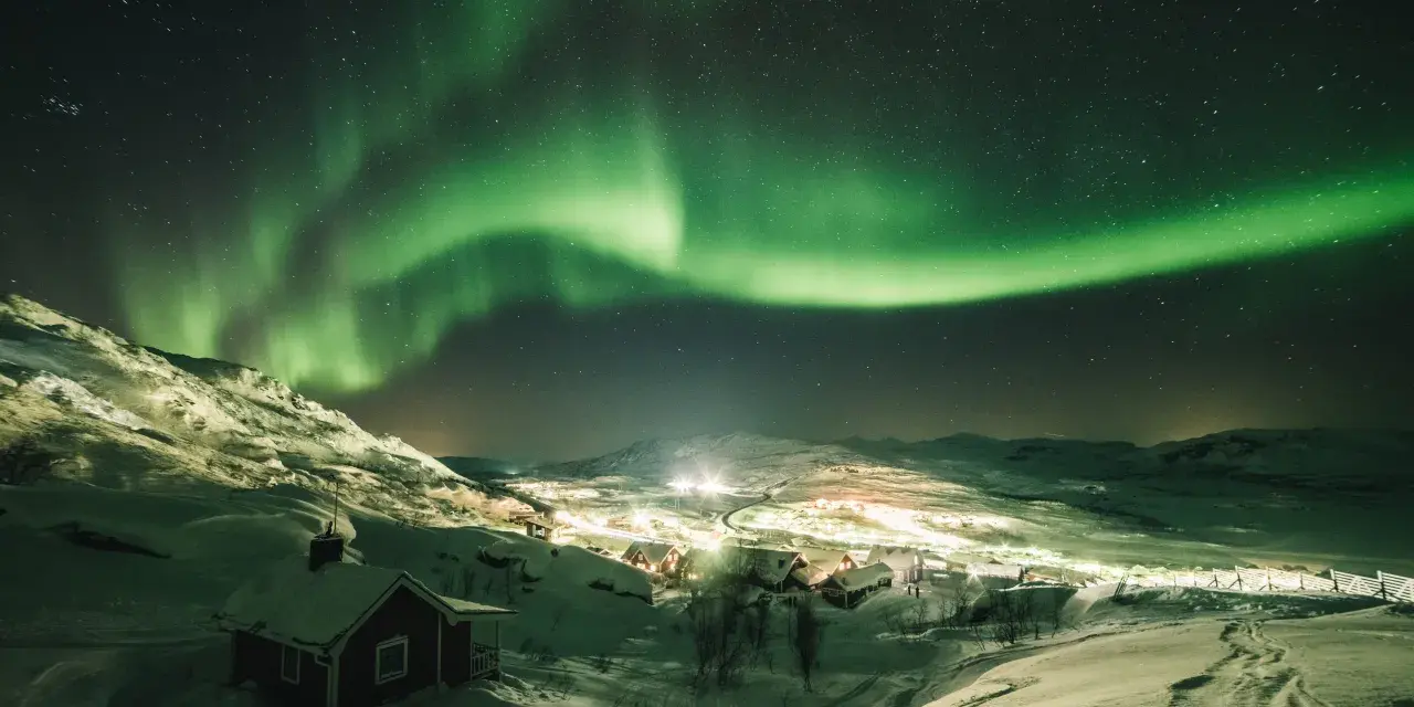 Snöklädda berg och en liten by badar i norrskenets gröna sken. En magisk natt att se norrsken.